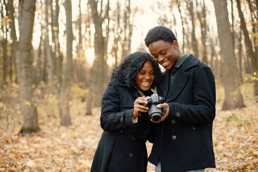 Black young man and his girlfriend holding a camera in hands. Romantic couple walking in autumn park. Man and woman looking on photo they made.